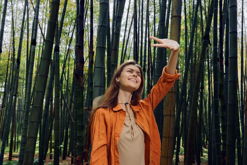 Woman bamboo forest portrait smile nature travel jacket: woman in orange jacket smiling and posing in a dense bamboo forest, portrait of travel and nature exploration with sunlight filtering.