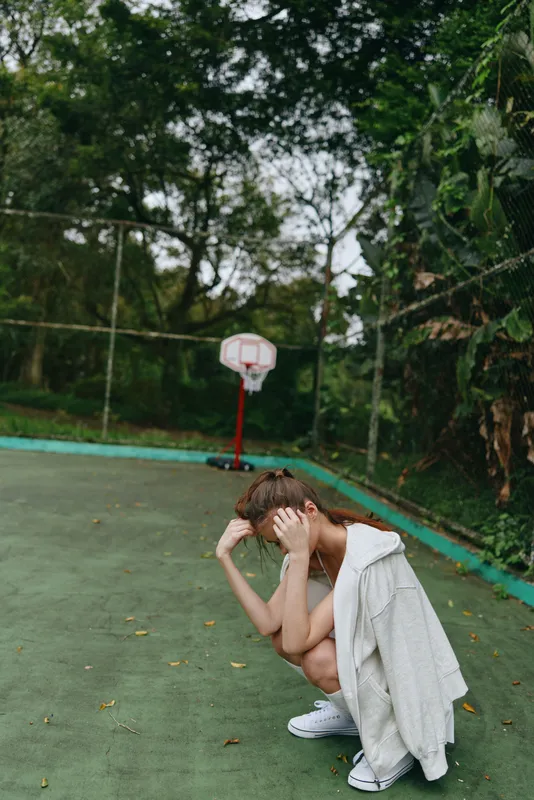 Young athletic model in sporty attire showing emotion on a basketball court, surrounded by greenery and soft natural light, conveying feelings of contemplation and relaxation