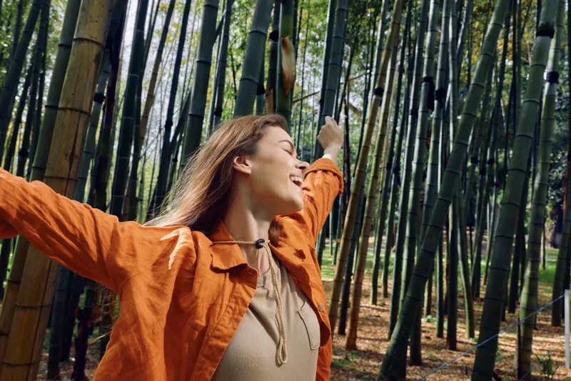 Woman in bamboo forest outdoors, portrait of happiness and freedom as she stretches arms, smiling in orange jacket, enjoying dappled sunlight and tranquil nature among tall green trees.