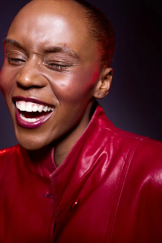 Close-up of joyful black woman with dark skin laughing with eyes closed wearing a red leather jacket. Studio portrait with vibrant mood and expressive emotion on purple background.