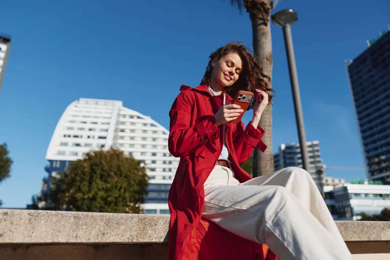 Young woman in a red coat sits on a concrete ledge outdoors while using her smartphone with a smile on a sunny day in the city.