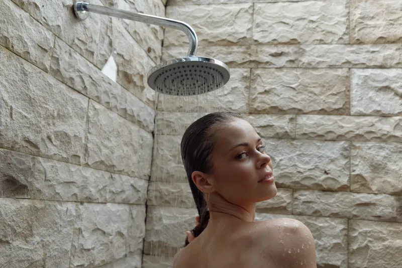 Woman taking an outdoor shower with stone wall background, wet hair, natural light, and refreshing water droplets creating a relaxing atmosphere.