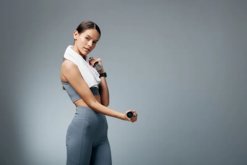 Woman fitness sport: Young fit woman in gray activewear holding small dumbbells with a white towel draped over her shoulders, posing confidently against a smooth neutral gray studio background.