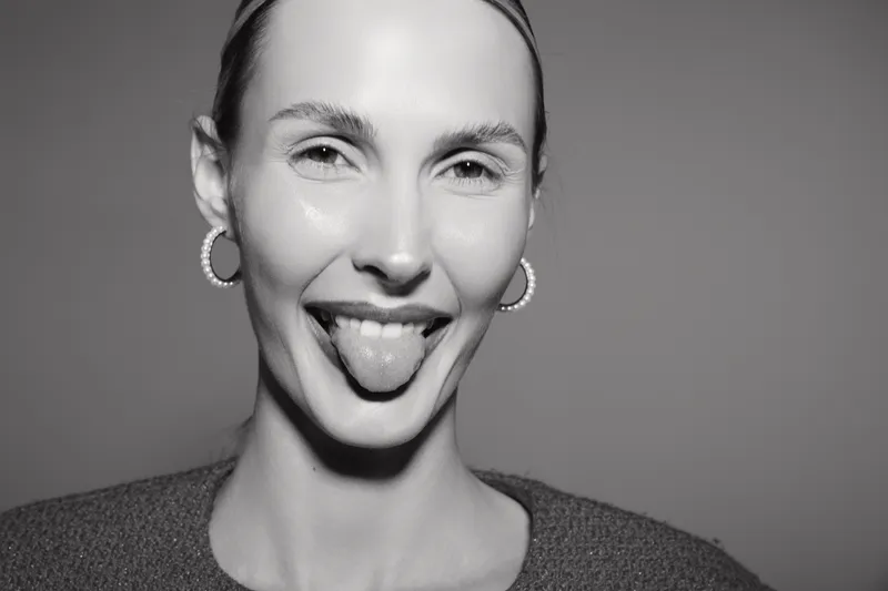 Playful young caucasian woman with fair skin and earrings sticks out tongue while smiling. Studio black and white close-up portrait with casual hairstyle and joyful expression.