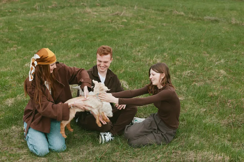 People outdoors on grass warmly interact with a goat kid, smiling and gentle. Casual lifestyle moment emphasizing bonding, nature, and animal companionship. Natural daylight creates a relaxed, joyful