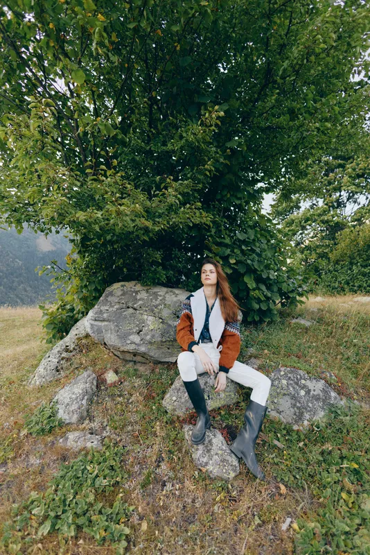 Woman sitting on rock under a large tree in countryside, wearing coat and boots, relaxed portrait in nature meadow with scarf and casual autumn fashion style.