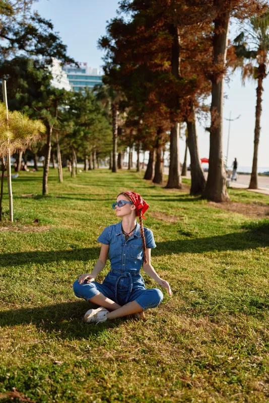 young woman meditating outdoors in casual denim outfit with sunglasses and red headscarf sitting cross-legged on grass under tall trees in sunny park