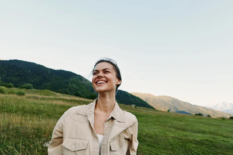 A happy woman in casual beige jacket smiles outdoors in a green field with mountains in the background under clear sky. Lifestyle concept of freedom and nature enjoyment.