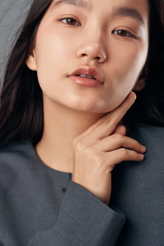 Young Asian woman with clear skin and natural makeup posing with hand near face for beauty portrait, fashion, elegance and skincare concept with soft lighting in studio setting.