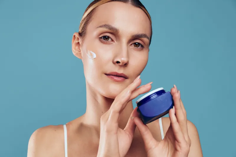 Woman skincare moisturizer cream facial beauty routine glow, hydration and healthy skin visible as she applies from a blue jar, close up studio portrait style, bright clean background
