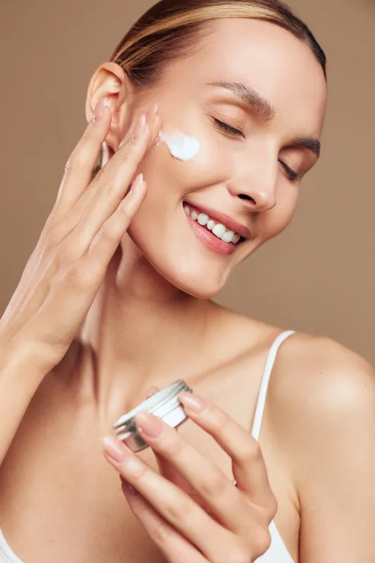 A smiling young woman applies white face cream on her cheek using fingers in a bright studio. Skincare and beauty concept with natural healthy skin and self care routine.