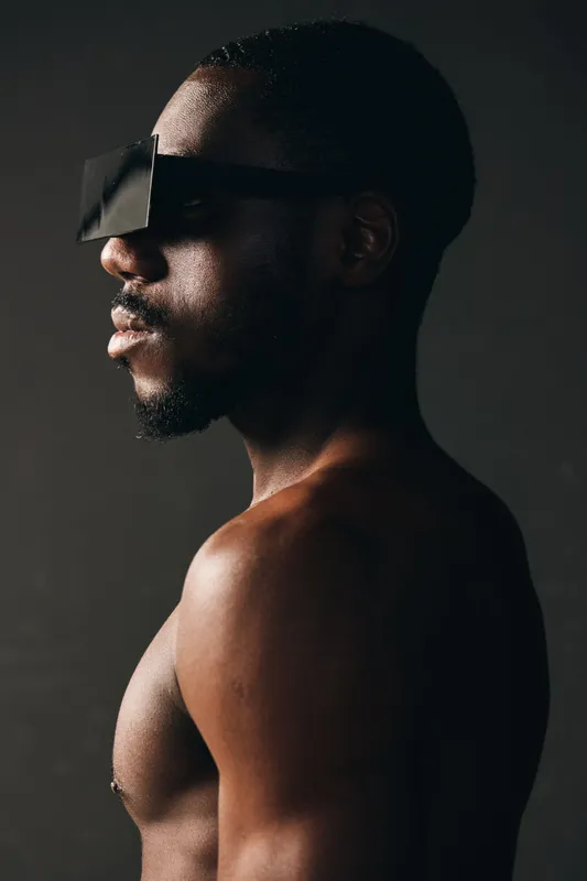 Creative conceptual portrait of African American man with futuristic black glasses in dark studio. Side view of shirtless male model with strong light and shadow contrast on skin.