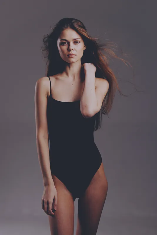 slender woman in black bodysuit posing with serious expression against gray background, stylish, minimalist