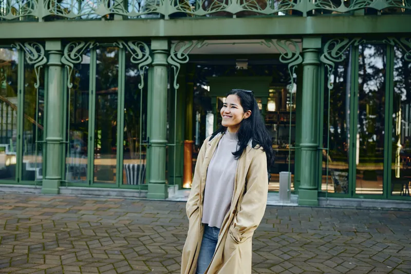 Woman in a beige coat outdoors, smiling beside a green decorative entrance, park backdrop, casual fashion vibe, candid mood, daylight and greenery