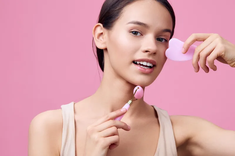 Young woman using skincare tools for facial massage on a pink background, promoting self care and beauty, highlighting a healthy skincare routine, and products for radiant skin