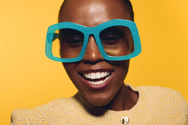 Happy African woman with dark skin wearing large blue sunglasses and a yellow sweater smiles brightly against yellow background. Concept for fashion, joy, and vibrant lifestyle.