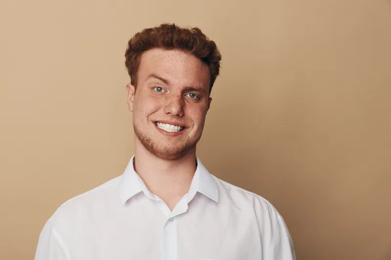 Smiling young man with curly hair and beard wearing white shirt stands against beige background. Portrait of confident male model with casual style and friendly expression.