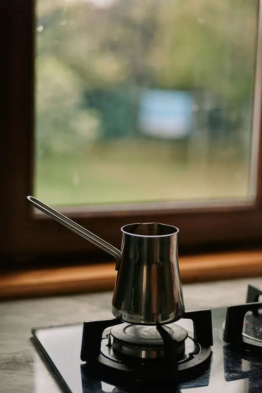 Closeup of a metal coffee pot on a stovetop burner beside a window, capturing a rustic kitchen moment of brewing coffee with warm light and soft steam
