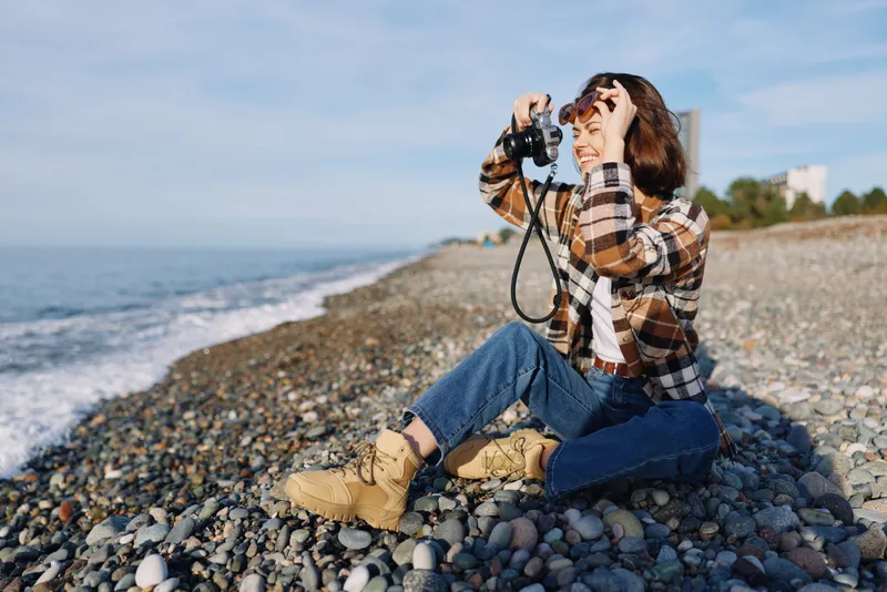 Photographer woman on a pebble beach captures the ocean using a camera, wearing a plaid shirt and boots, seated by the shoreline, enjoying nature and outdoor photography.