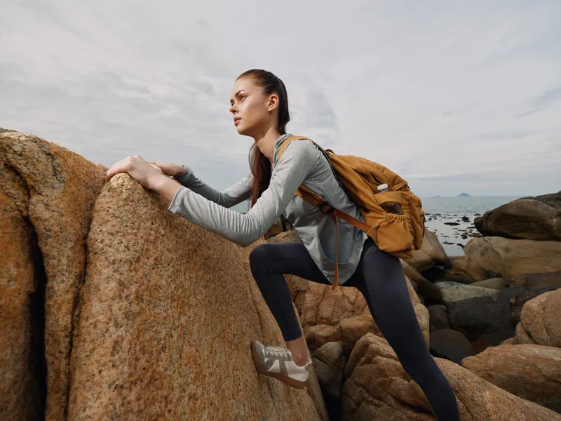 Young female hiker climbing rocks, wearing sporty attire with a backpack, showcasing determination against a coastal background, perfect for adventure themed content