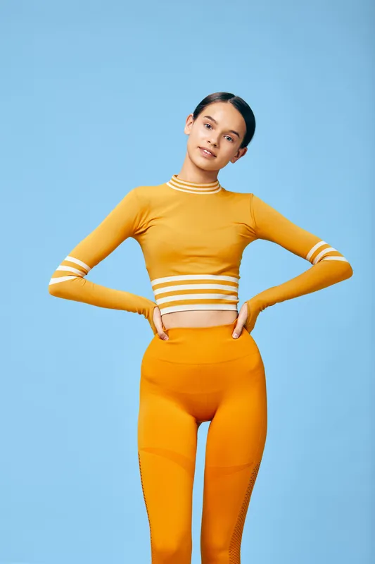 Woman fitness model in bright orange athletic wear poses confidently against a blue studio backdrop, highlighting athleticism, balance and a healthy active lifestyle.