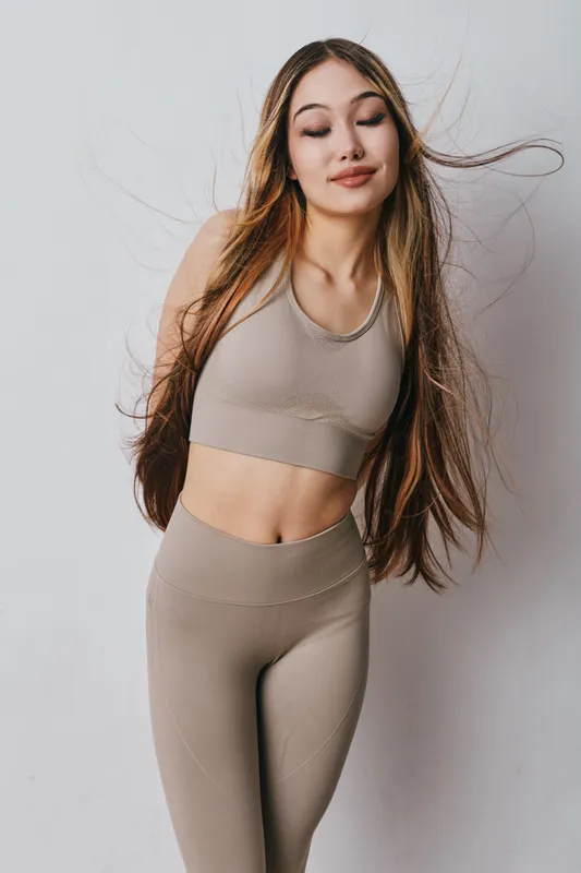 Young woman with long hair wearing seamless beige sportswear stands with closed eyes against white background for fitness, health, and lifestyle concept.