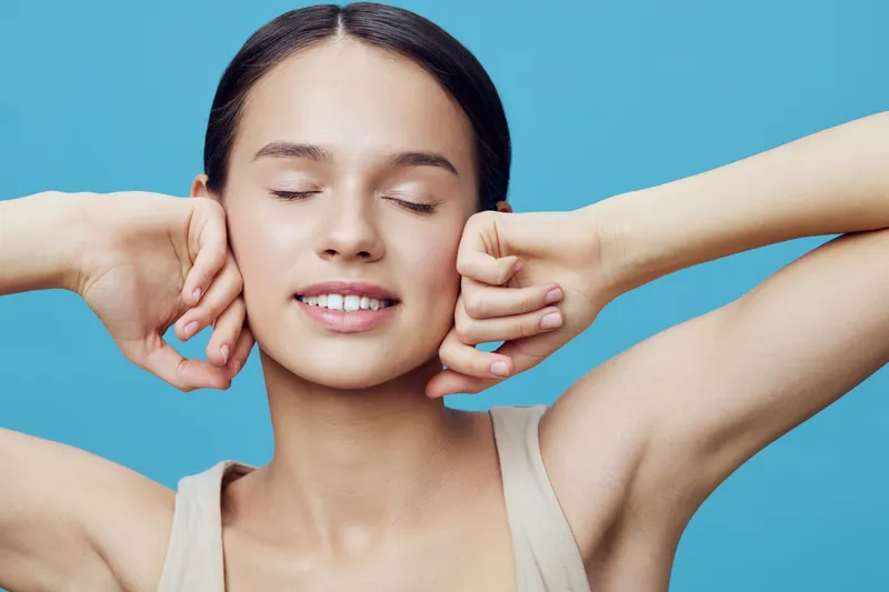 Young woman enjoying a moment of relaxation with closed eyes, wearing a neutral tank top against a soft blue background Her serene expression conveys tranquility and joy