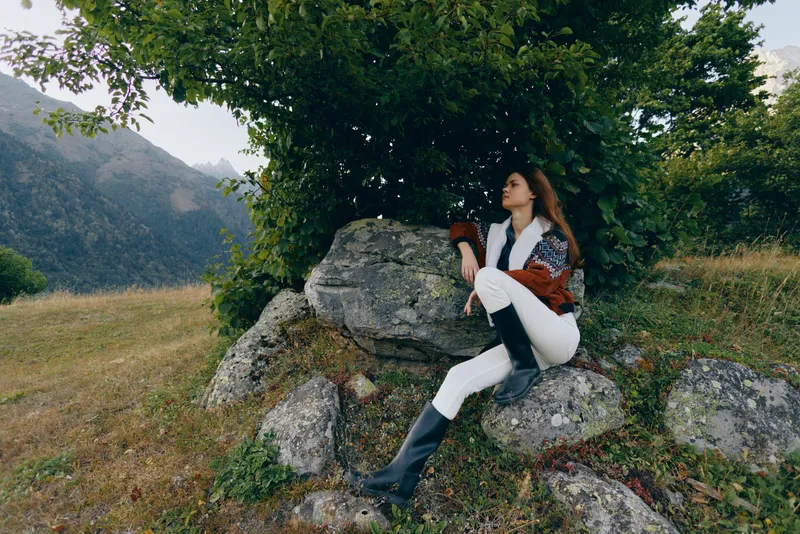 Woman sitting on rocks under a tree in a mountain meadow wearing boots and a jacket, relaxed portrait with scenic mountains backdrop and natural outdoor lifestyle atmosphere.