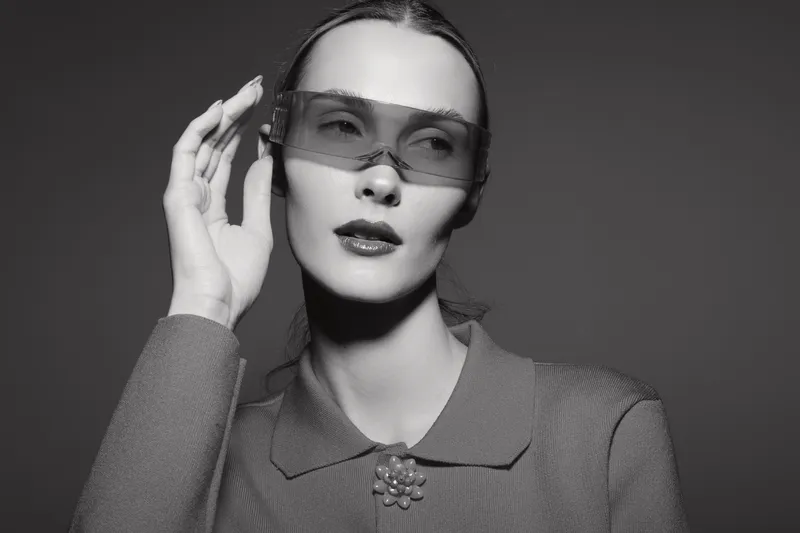 Young white woman with pale skin wears large futuristic sunglasses and vintage outfit, posing with hand near face in monochrome studio portrait.