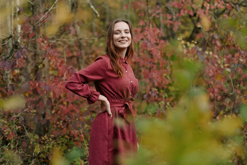 A candid portrait of a smiling woman in a burgundy dress amid autumn foliage, conveying authenticity, credible mood, warm light, and natural charm.