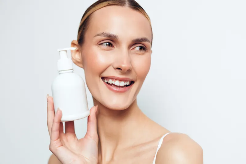 Smiling woman holding face cream bottle near cheek, closeup on white background, smooth skin beauty care concept, soft studio lighting for wellness and confidence