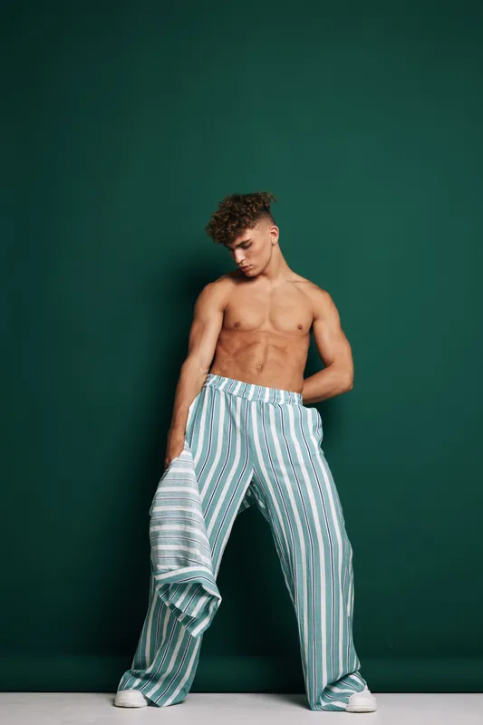Young man, shirtless and wearing striped wide pants, posing against dark green background. Male fashion model, trunk bare with casual relaxed modern style in studio shot, indoor lifestyle.