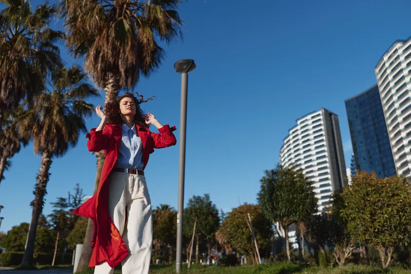 Young woman in a red coat walks outdoors in a sunny city park with palm trees and modern buildings in the background