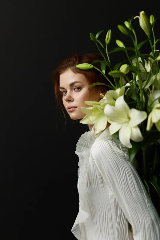 A young woman with a serene expression holding white lilies, dressed in a flowing white blouse, against a dark background, symbolizing beauty and tranquility