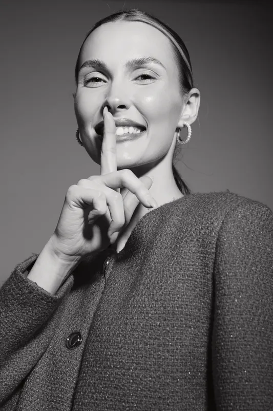 Happy Caucasian woman with fair skin smiling and making silence gesture with finger on lips. Close-up portrait in textured sweater with hoop earrings on gray background. Concept of secrecy.