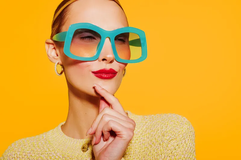 Stylish woman with large turquoise sunglasses and red lipstick poses thoughtfully on bright yellow background. Fashion concept with colorful accessories and confident expression.