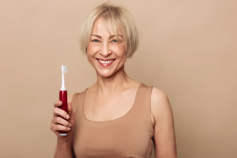 Smiling mature woman with short blonde hair holds electric toothbrush on beige background. Concept of dental care, oral hygiene, healthy lifestyle and personal grooming routine.