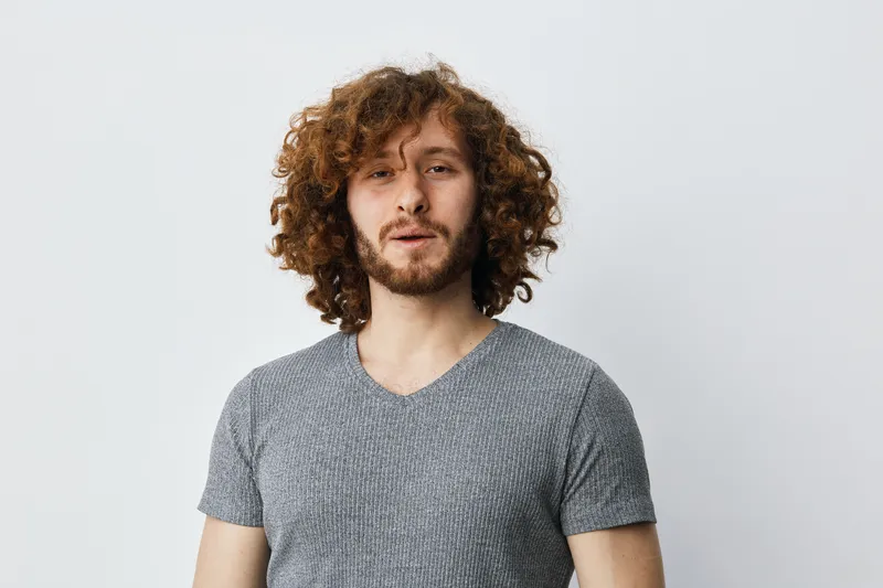 Portrait of a young man with curly hair smiling in a casual gray t shirt against a soft white background, conveying confidence and approachability