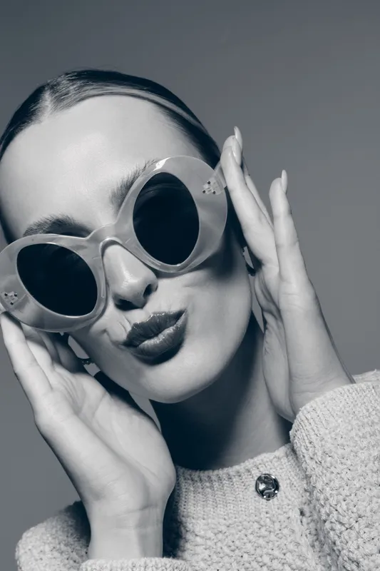 Young Caucasian woman with light skin wearing oversized round sunglasses and a knitted sweater poses confidently on a plain background. Fashion and style concept.