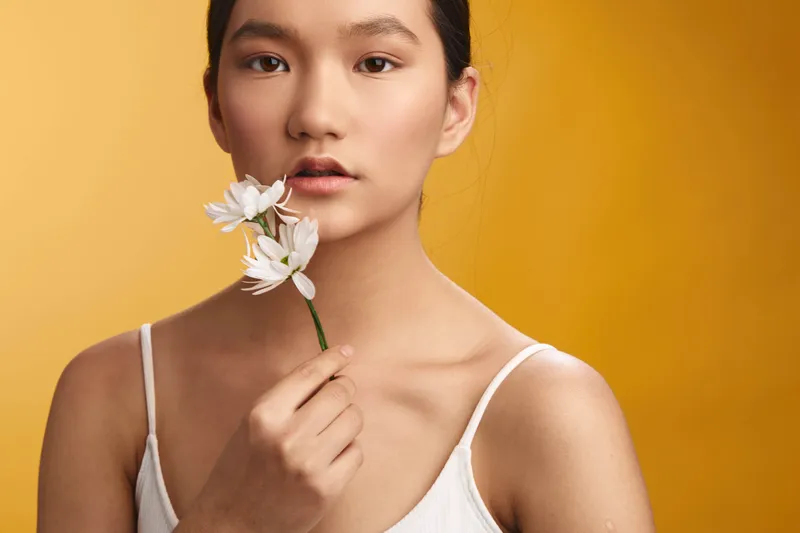 Asian woman is captured in a studio portrait holding a delicate flower near her lips against a warm yellow backdrop, conveying grace and calm in a minimalist setting.
