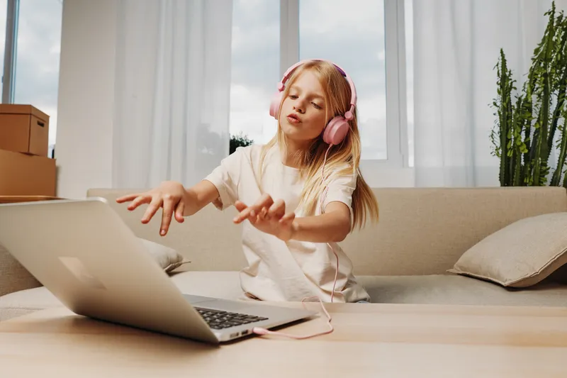 Blonde girl with pink headphones enjoying music while using laptop in bright living room. Concept of childhood, technology, and leisure time at home.