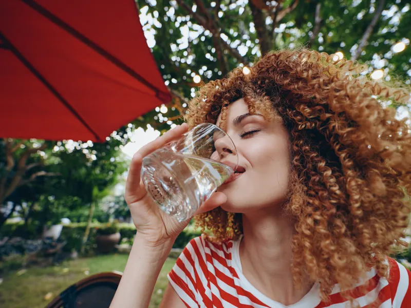 Portrait of a woman with curly hair drinking water from a glass outdoors under a red parasol, enjoying a refreshing moment in a sunny garden setting.