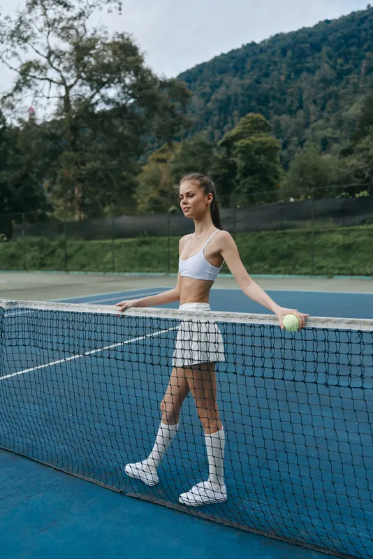 Young female tennis player in athletic wear poses confidently by the net on a tennis court, embodying strength and focus during outdoor sports activity