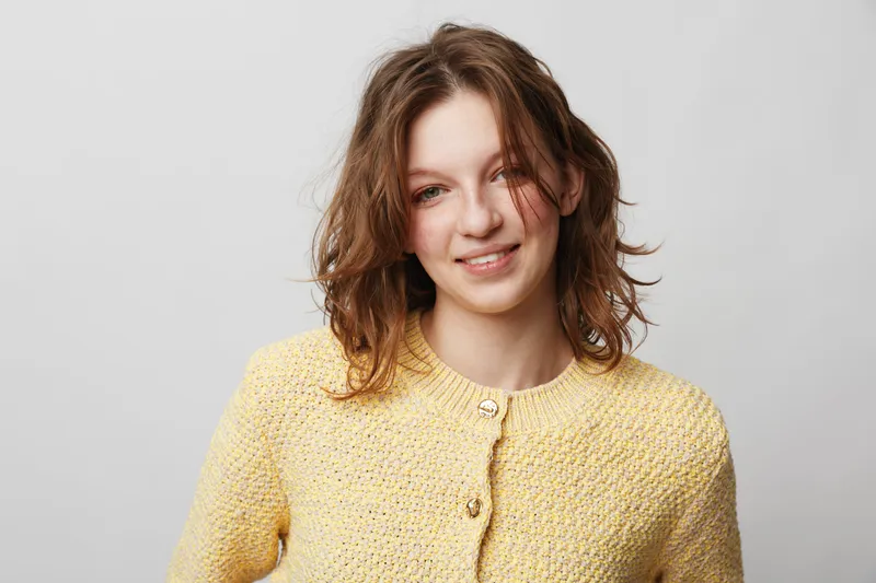 Smiling young woman with wavy brown hair wearing a yellow knitted sweater stands against a plain light background. Concept of casual style, happiness, and natural beauty.