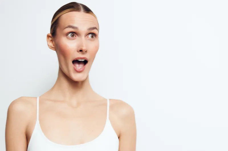 Woman closeup applying face cream for skincare wellness and selfcare, surprised expression, studio light on clean background. White sleeveless top signals fresh hygiene routine and healthy complexion.