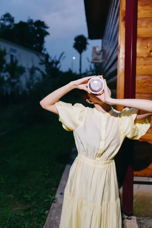 Outdoor scene with a person in a yellow dress holding a round object up to the face, creating a playful casual portrait at dusk with soft dusk light.
