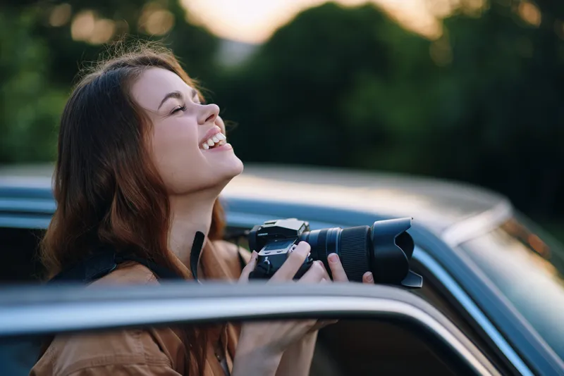 Cheerful young woman photographing nature with a camera, showcasing happiness and joy in a serene outdoor setting during sunset.