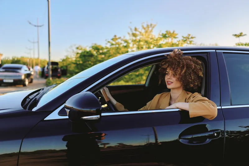 Woman with curly hair sits in a driver seat of a car, embracing a lifestyle moment with retro film color, sunlight on highway, casual stylish mood, natural light and confidence