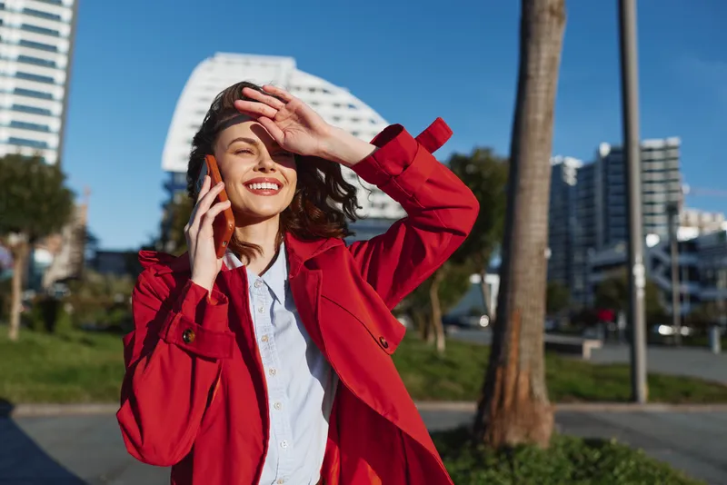 Young woman in a red coat talks on a phone outdoors in an urban plaza, smiling as bright sunlight lights modern buildings and palm trees in the background, creating a lively street scene.