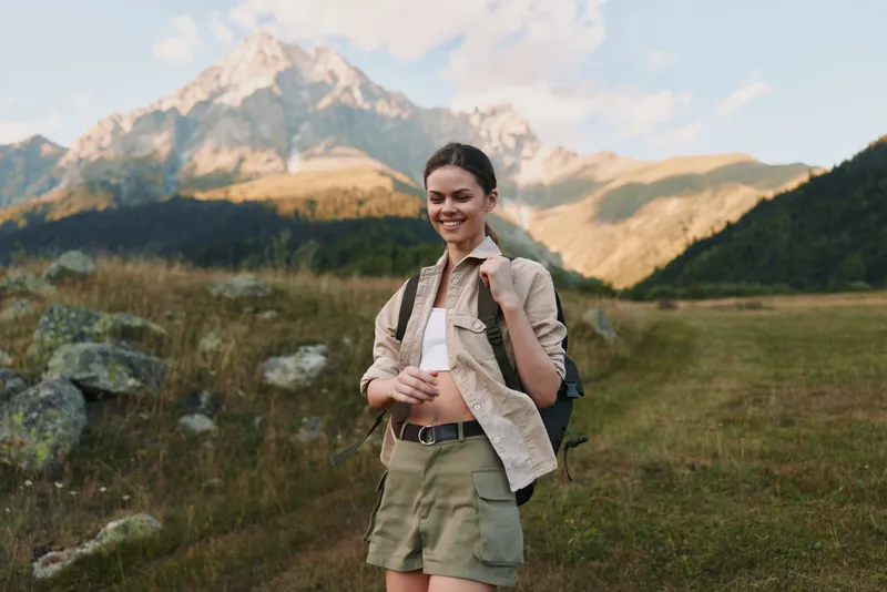 Young woman traveler with backpack stands in mountain landscape smiling enjoys nature adventure in sunny day wearing casual clothes and shorts in open field with rocky hills.
